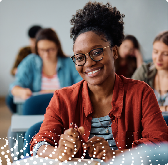 A smiling woman is sitting at a desk in a classroom setting. There are other people sitting at desks behind her.
