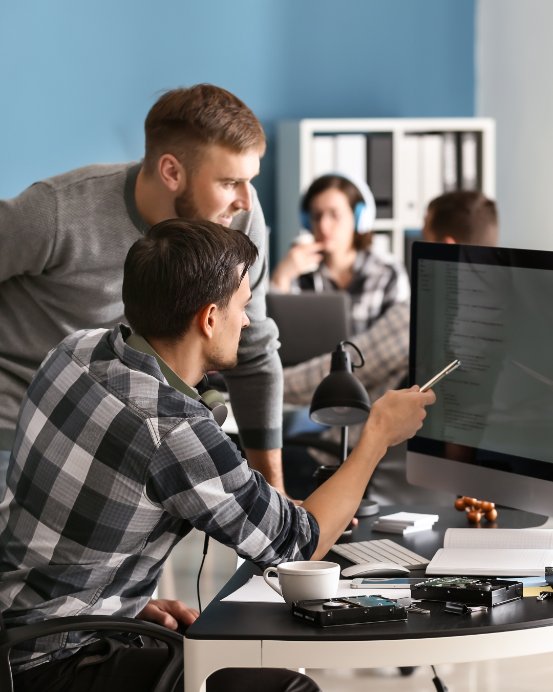 Two men at a desk working on a computer in an office setting. There are another man and woman at another desk in the background.